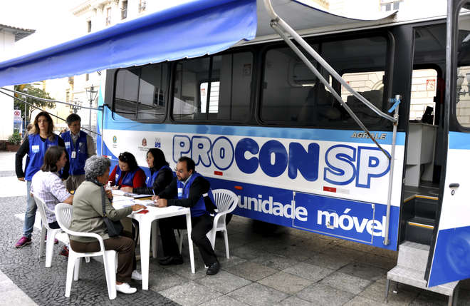 Foto mostra duas mulheres sendo atendidas por três funcionários do Procon-SP que estão uniformizados com um colete azul, todos sentados à mesa. Ao lado deles mais duas funcionárias acompanham o atendimento. No fundo da imagem se vê o ônibus do Procon-SP.
